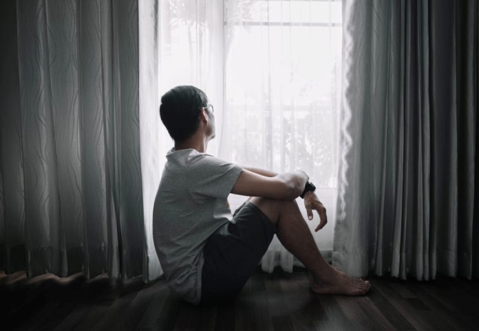 Sad and depressed young male sitting on the floor in the living room looking outside the doors,sad mood,feel tired, lonely and unhappy concept. Selective focus.